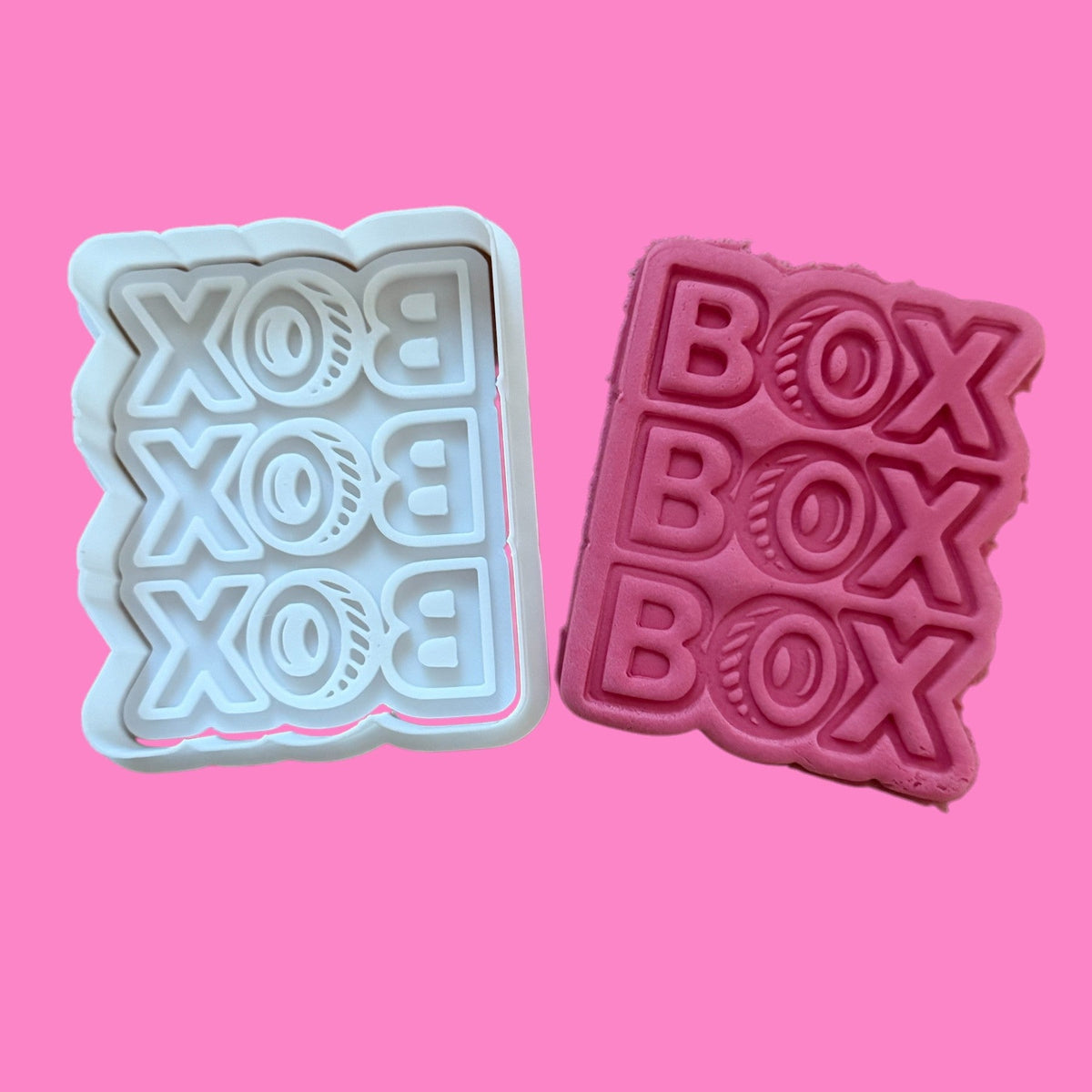 Two cookie cutters with a 'Box Box Box' racing-inspired design, one in white and one in pink, displayed against a pink background.