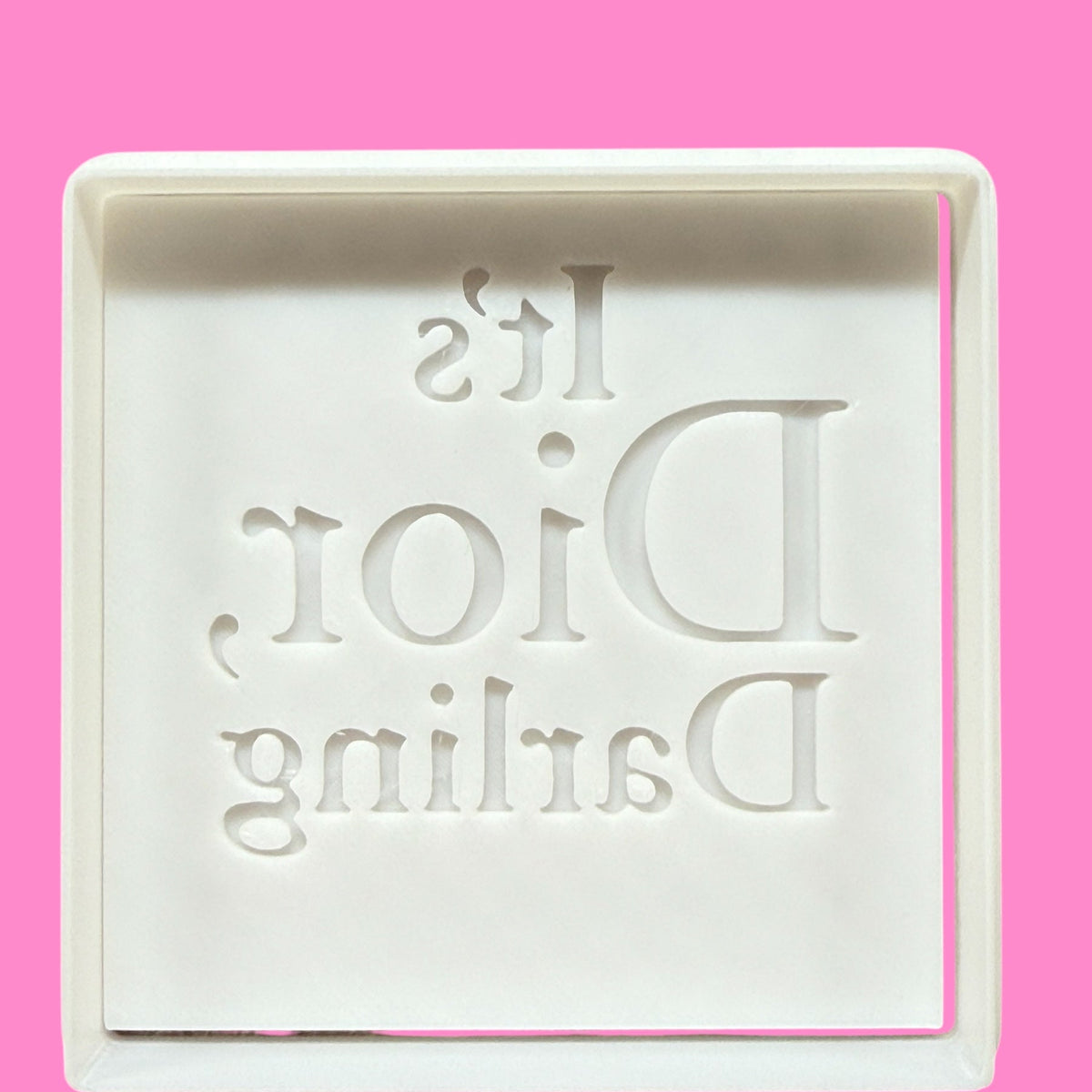 A plastic cookie cutter and embosser with the text 'It's Dior, Darling' engraved on it, against a pink background.