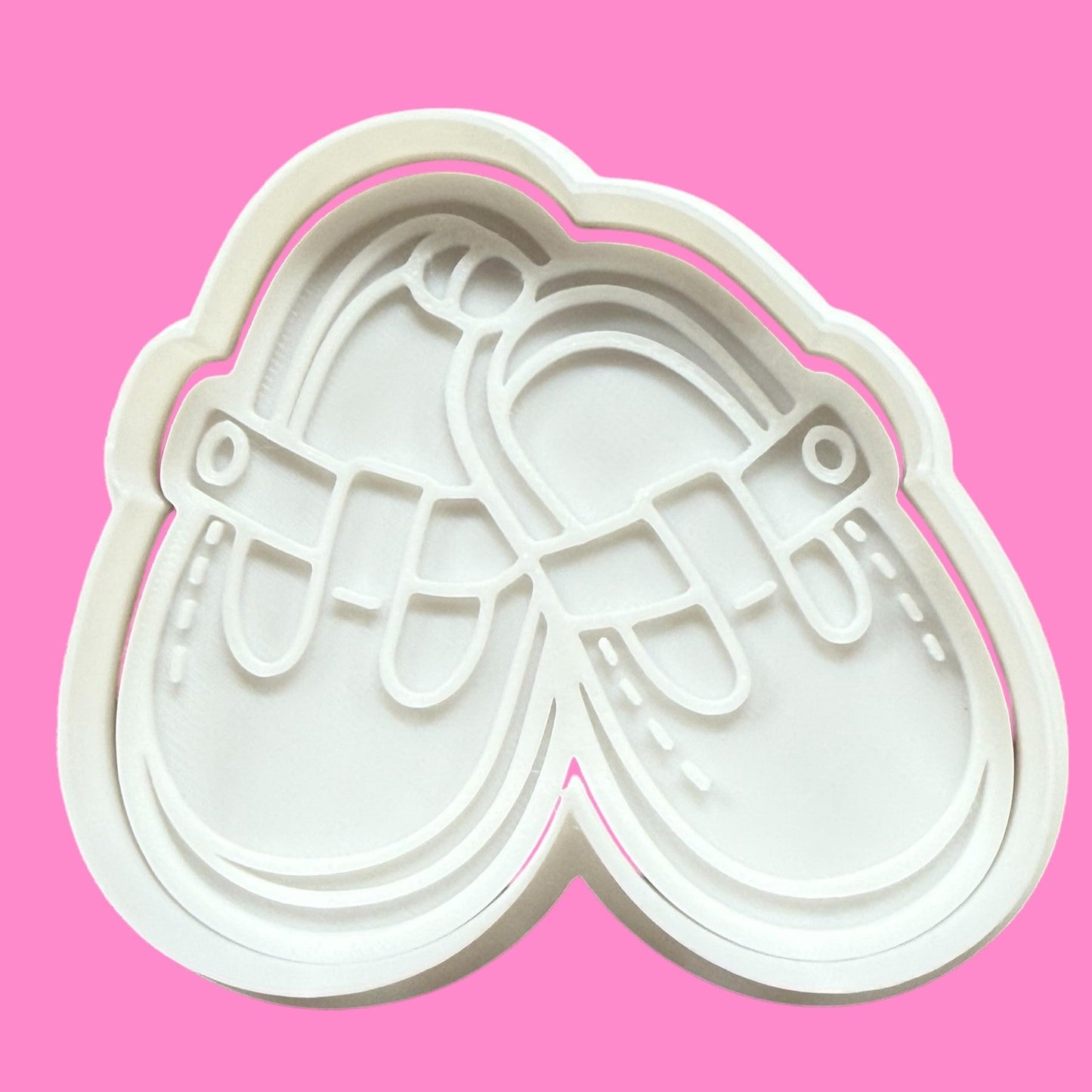 A white plastic cookie cutter and embosser in the shape of a pair of newborn baby shoes, displayed against a pink background.
