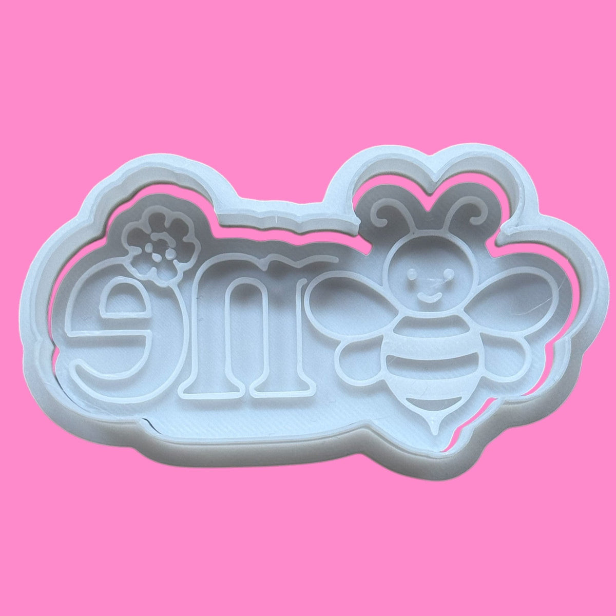 A plastic cookie cutter and embosser with a design that includes the number 1 and a bee, on a pink background.