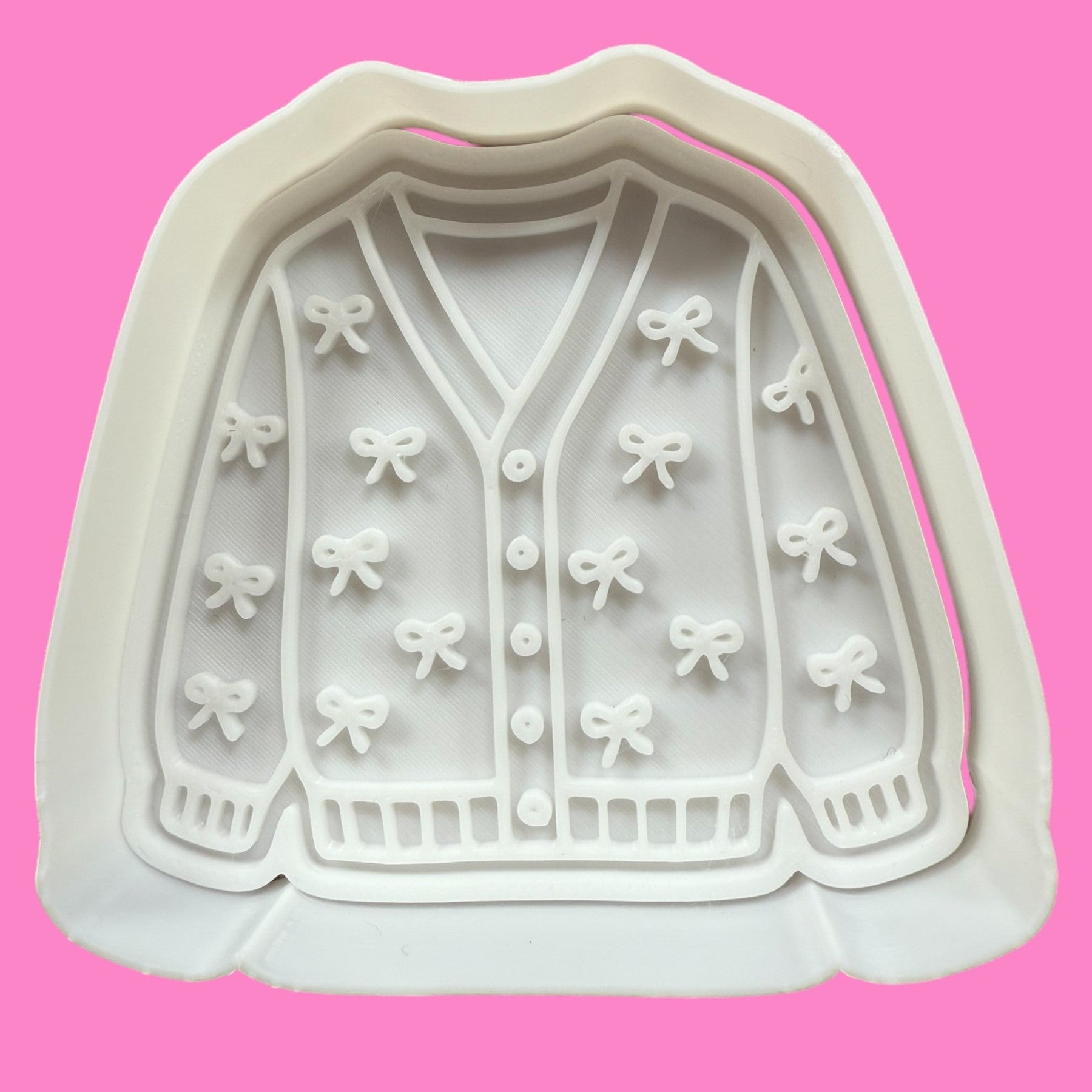 A cookie cutter and embosser with a cozy knit-inspired design, featuring cardigan texture with tied ribbon accents.