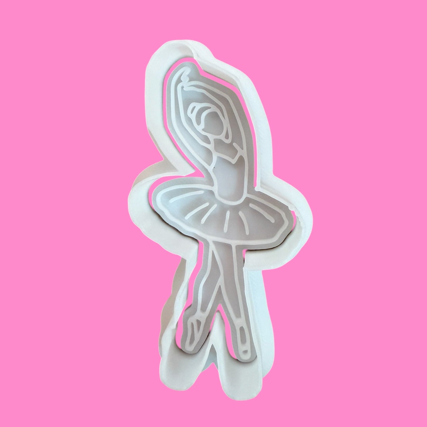 A cookie cutter and embosser in the shape of a classic ballerina silhouette, with fine details such as a tutu and hair bun, against a pink background.