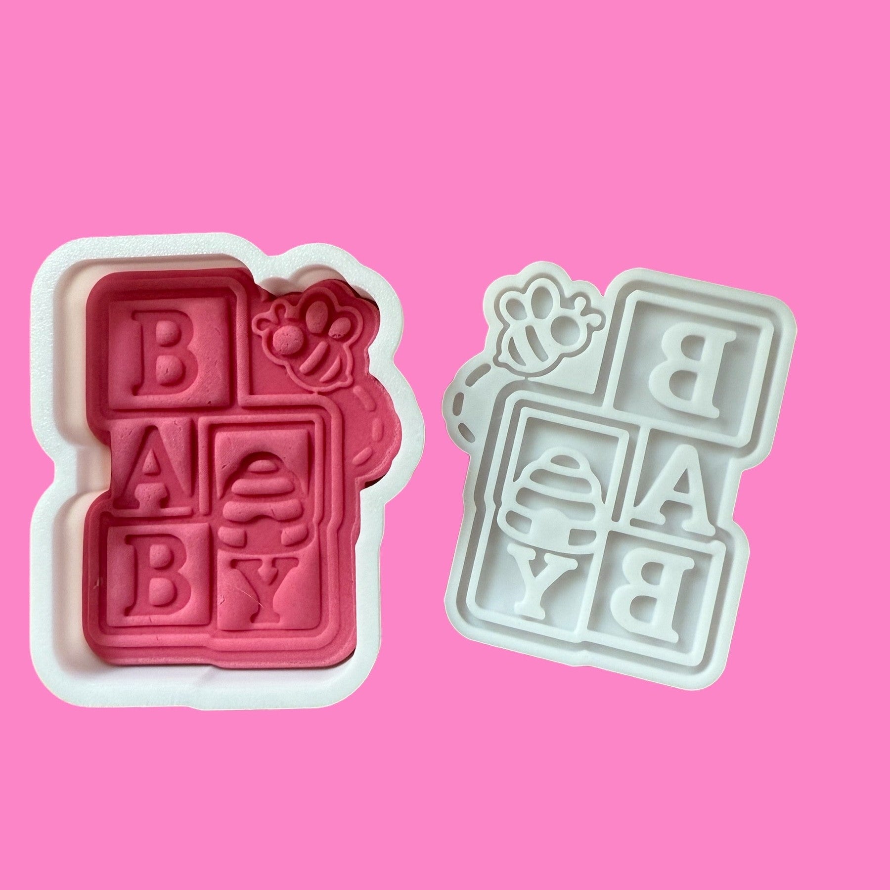 Two plastic cookie cutters in the shape of stacked baby blocks with a bumblebee design, one in red and one in white, displayed on a pink background.