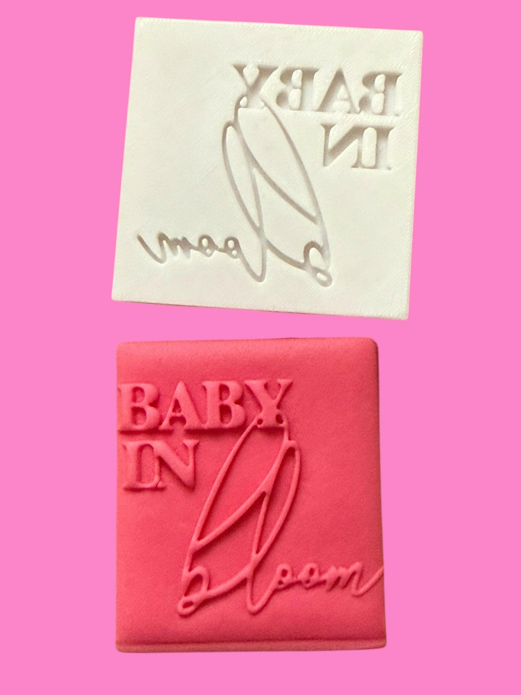 A pink square cookie cutter with a floral 'BABY IN BLOOM' design, accompanied by a matching font stamp.