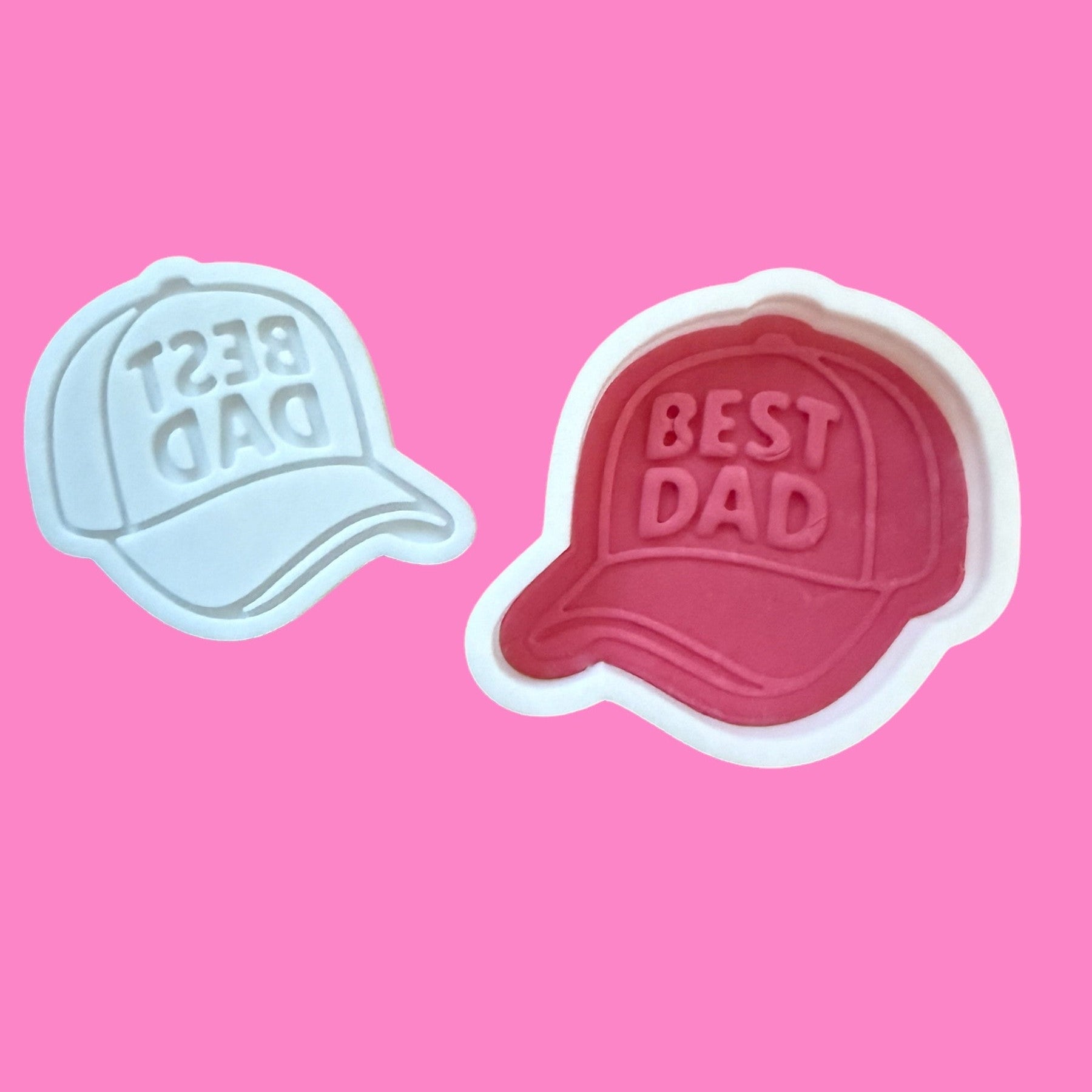 Two cookie cutters in the shape of baseball caps with 'BEST DAD' text embossed on them, one in red and the other in blue.