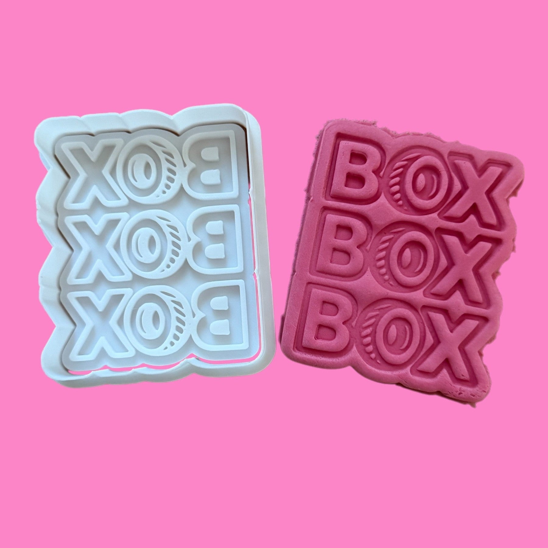 Two cookie cutters with a 'Box Box Box' racing-inspired design, one in white and one in pink, displayed against a pink background.