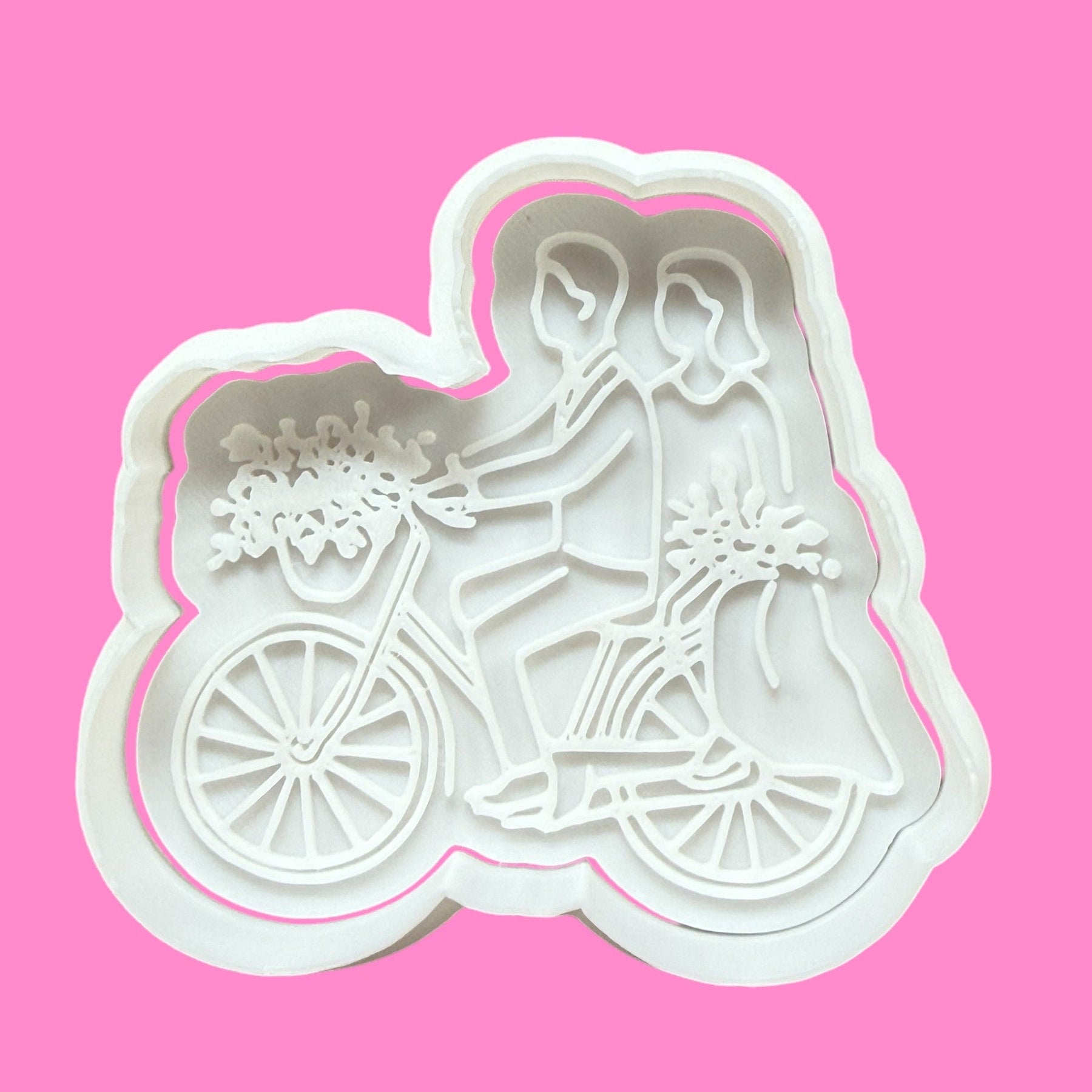 A white cookie cutter and embosser in the shape of a bride and groom riding a push bike with floral embellishments.