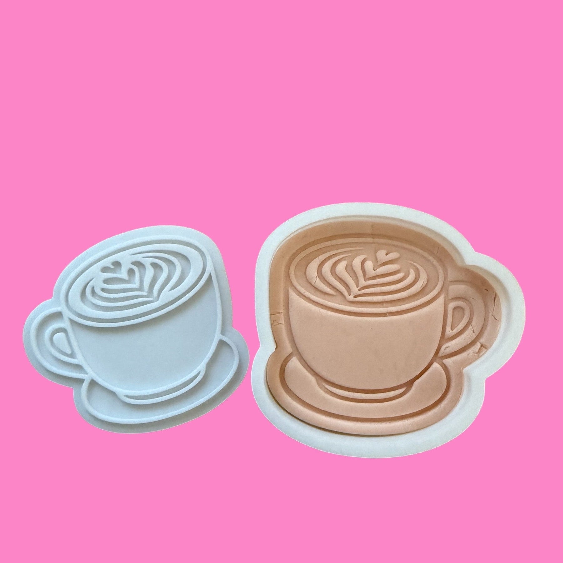 Two cookie cutters in the shape of coffee cups with latte art designs, one embossed and one cut-out, displayed against a pink background.