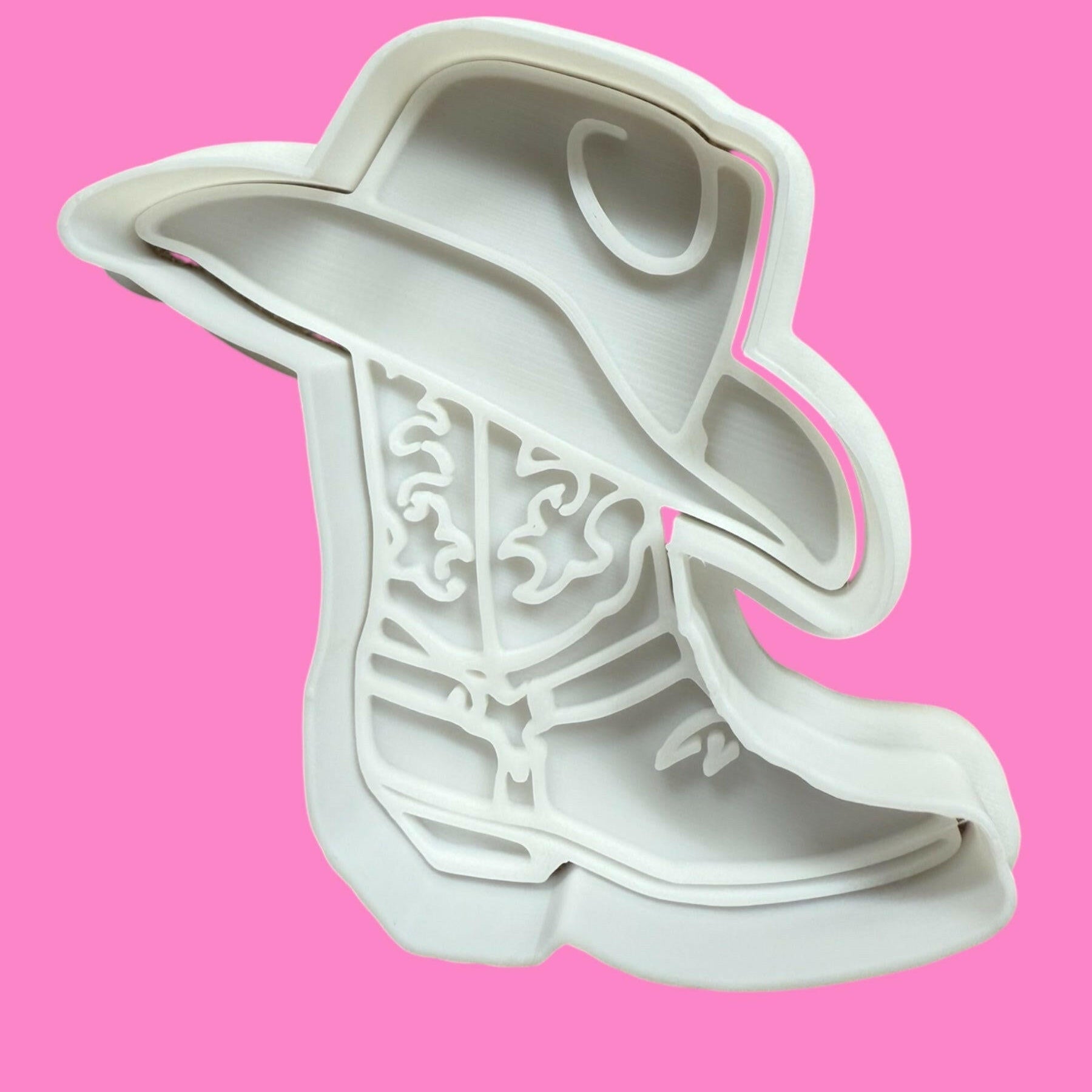 A white plastic cowboy hat on a cowboy boot cookie cutter and stamp set against a pink background, designed for western theme baking.