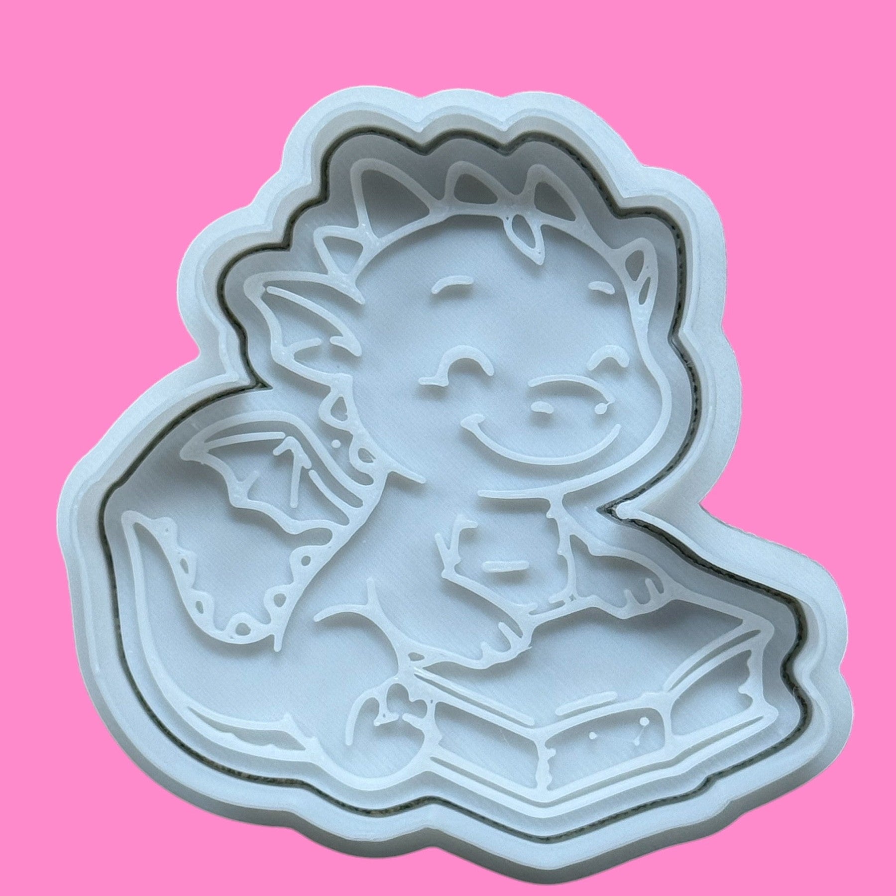 A plastic cookie cutter and embosser in the shape of a baby dinosaur sitting and reading a book, with a pink background.