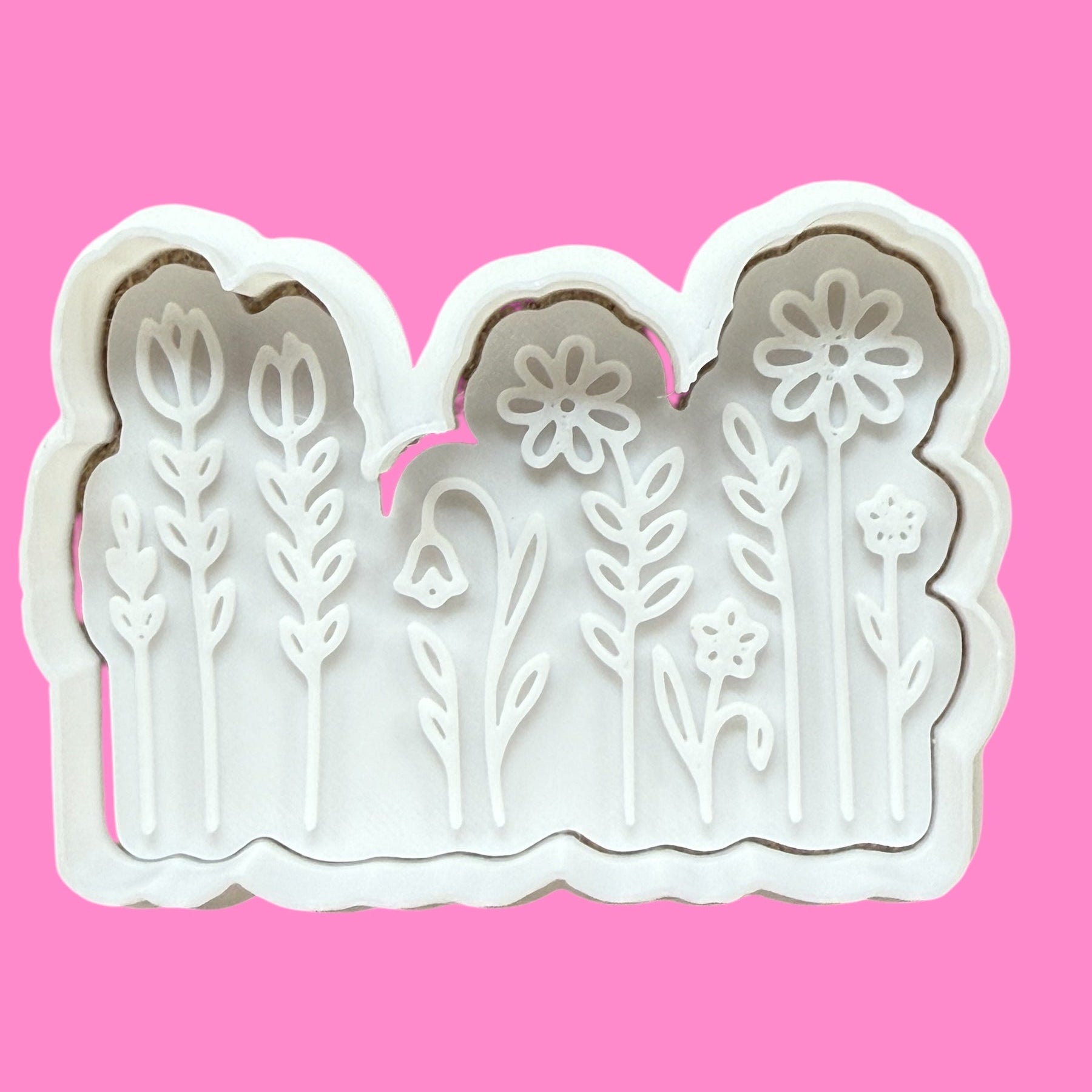 A cookie cutter and embosser set with floral spring flower designs, with intricate details resembling petals, leaves and stems, displayed against a pink background.