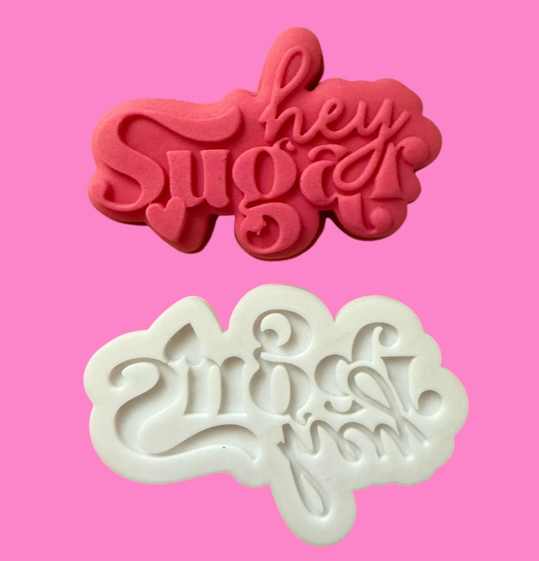 Two cookie cutters with the phrase 'hey sugar' and a heart shape, one red and one white, on a pink background.