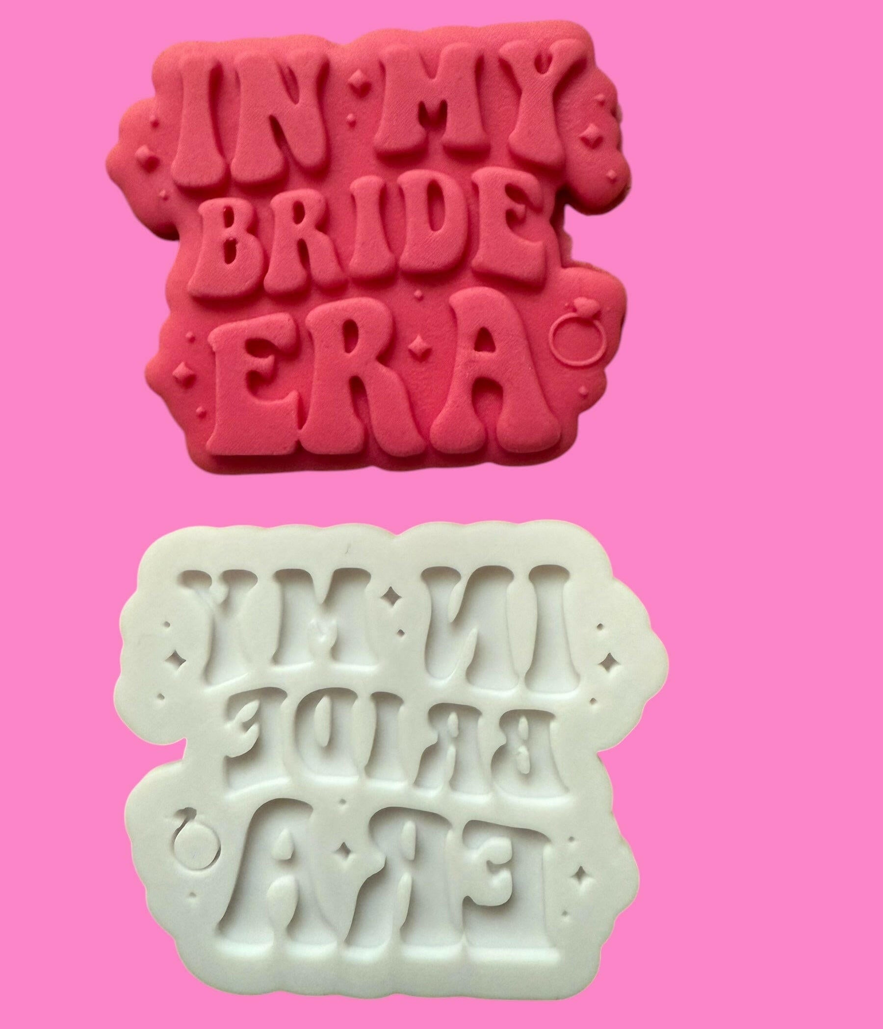 Two cookie cutters with the phrase 'IN MY BRIDE ERA' printed in a trendy font, one in red and one in white, against a pink background.
