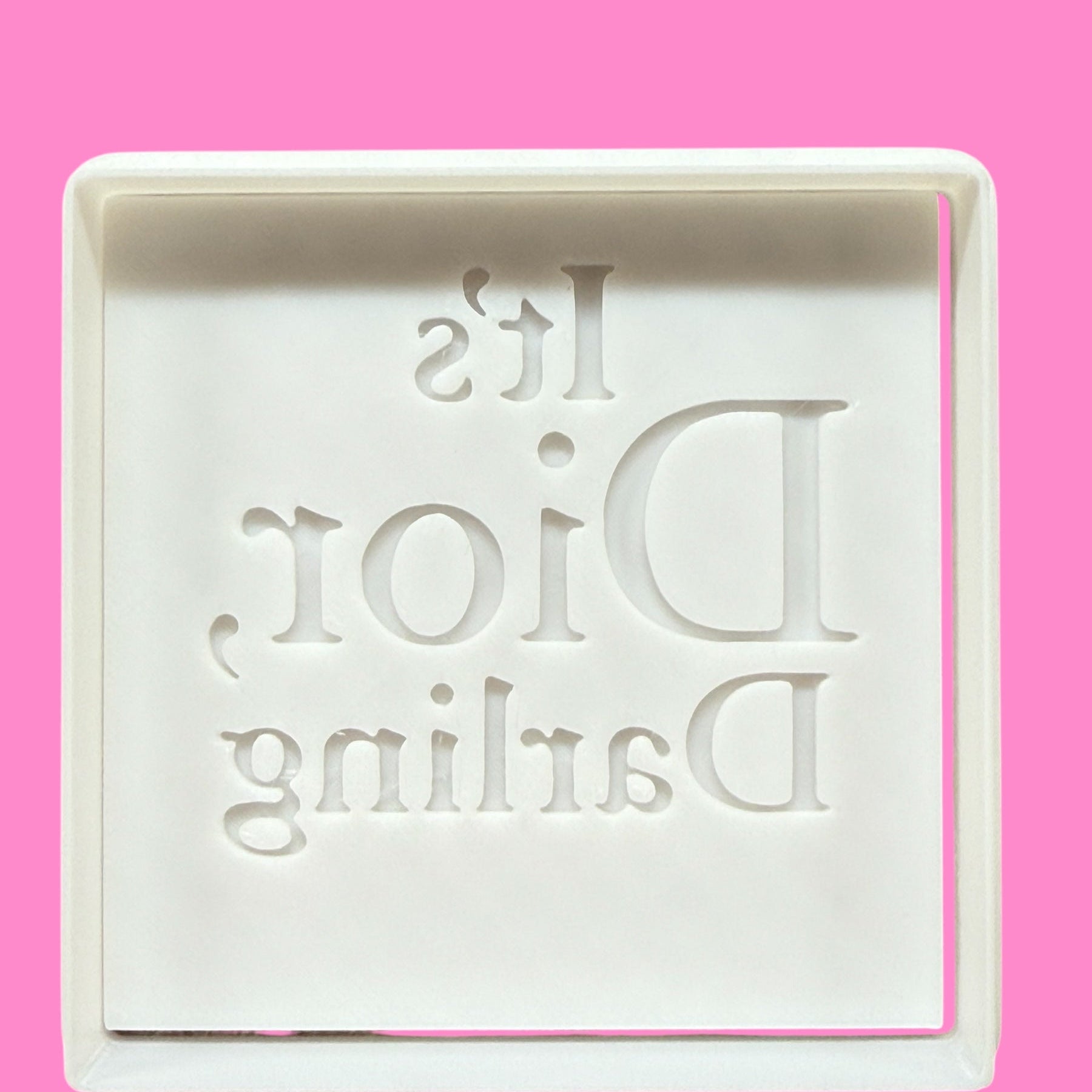 A plastic cookie cutter and embosser with the text 'It's Dior, Darling' engraved on it, against a pink background.