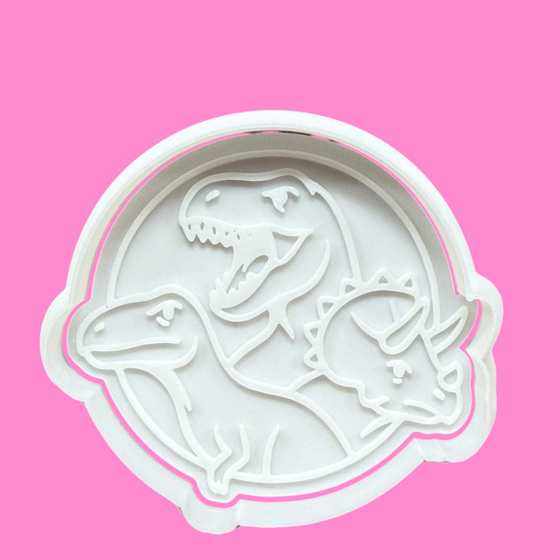 A white dinosaur-shaped cookie cutter and embosser on a pink background.
