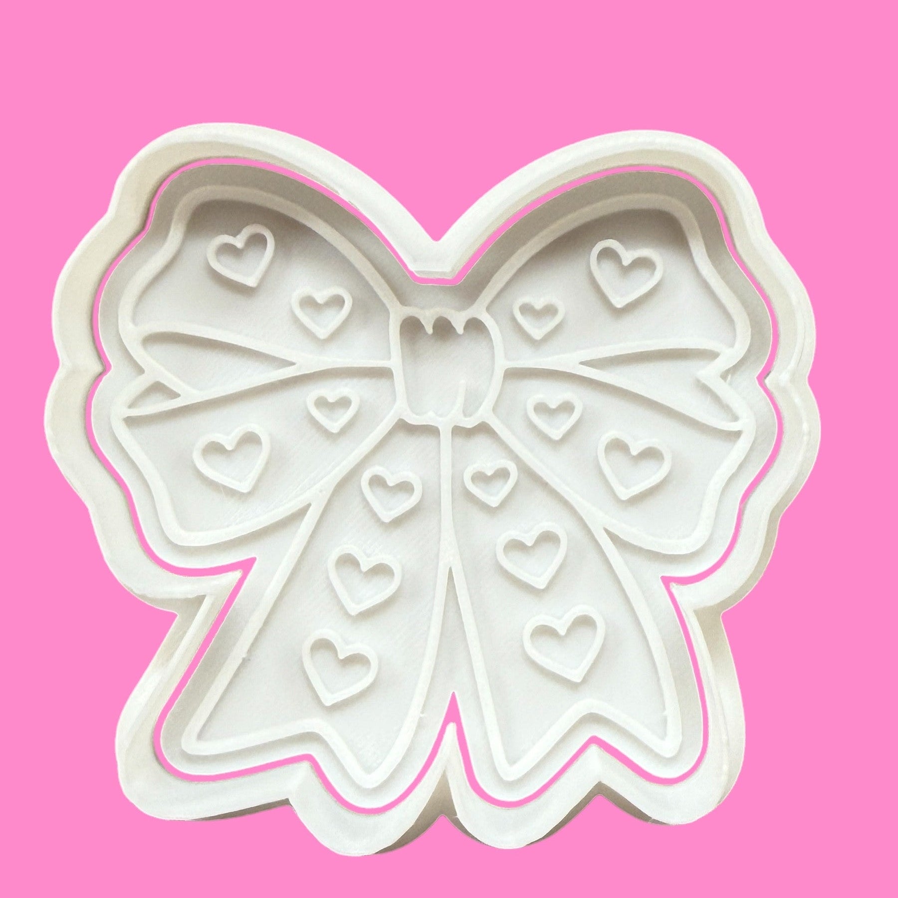 A white plastic cookie cutter and embosser in the shape of a ribbon bow with heart details on a pink background.