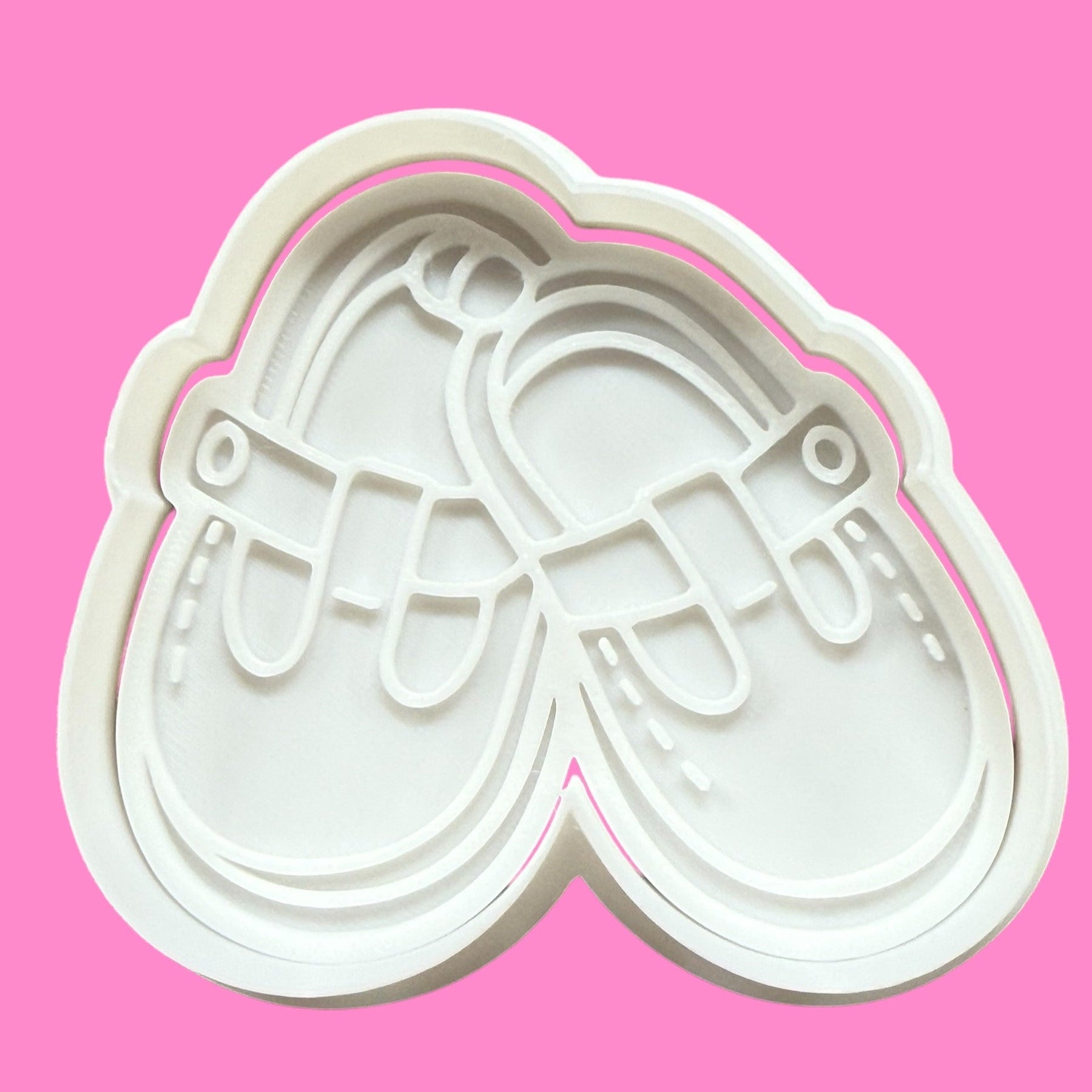 A white plastic cookie cutter and embosser in the shape of a pair of newborn baby shoes, displayed against a pink background.