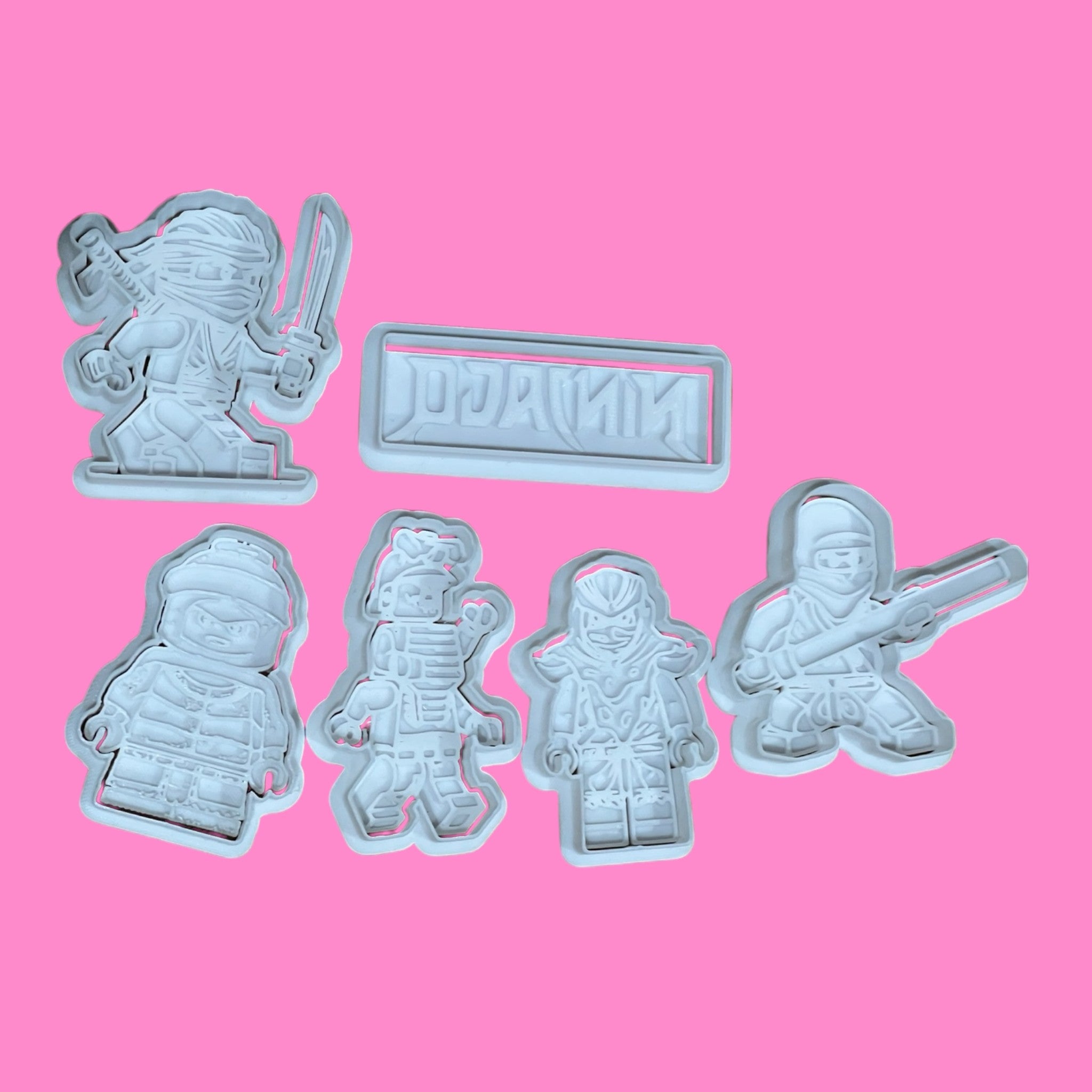 A collection of six ninja-themed cookie cutters and embossers, with various designs including ninja characters and a 'NINJAGO' logo, displayed on a pink background.