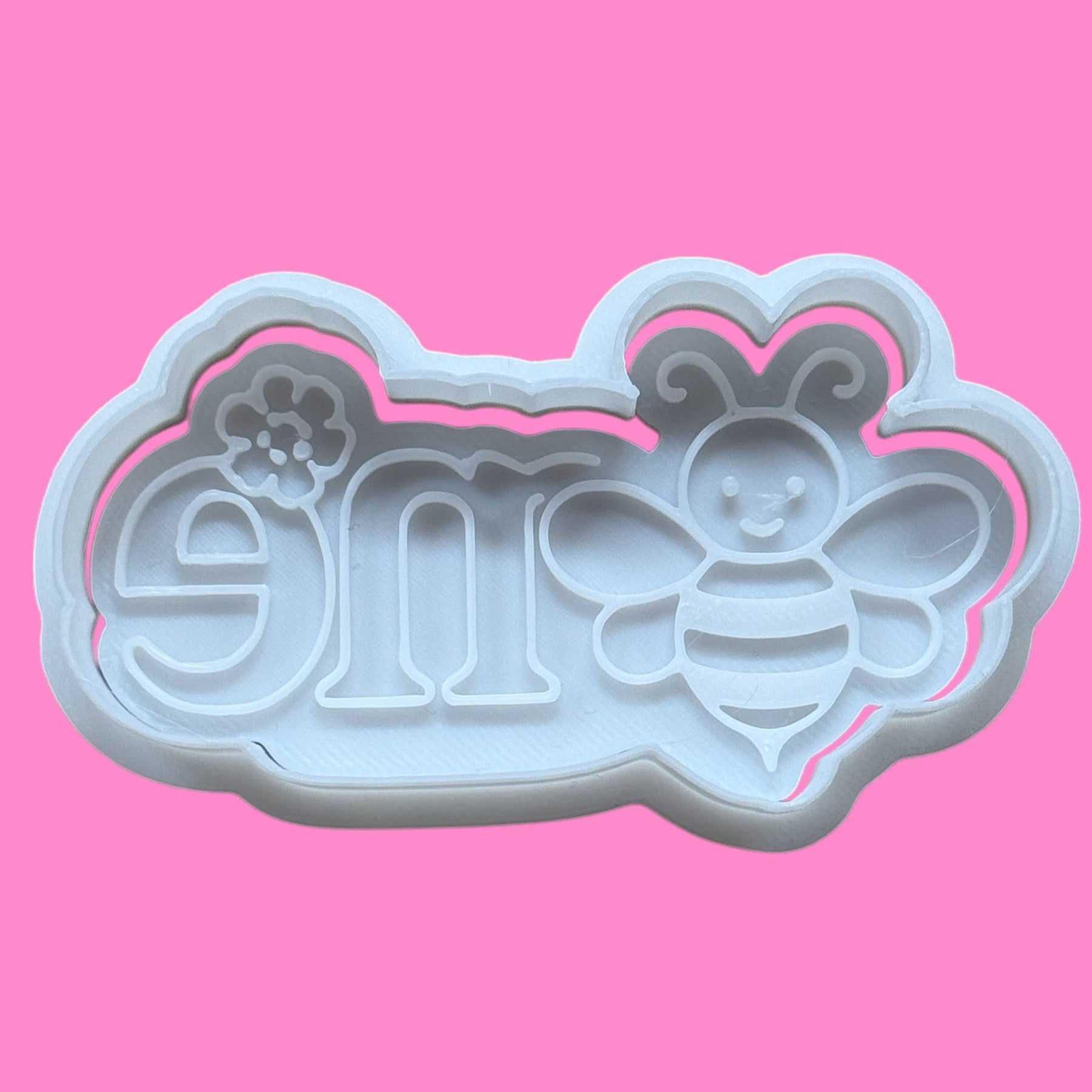 A plastic cookie cutter and embosser with a design that includes the number 1 and a bee, on a pink background.