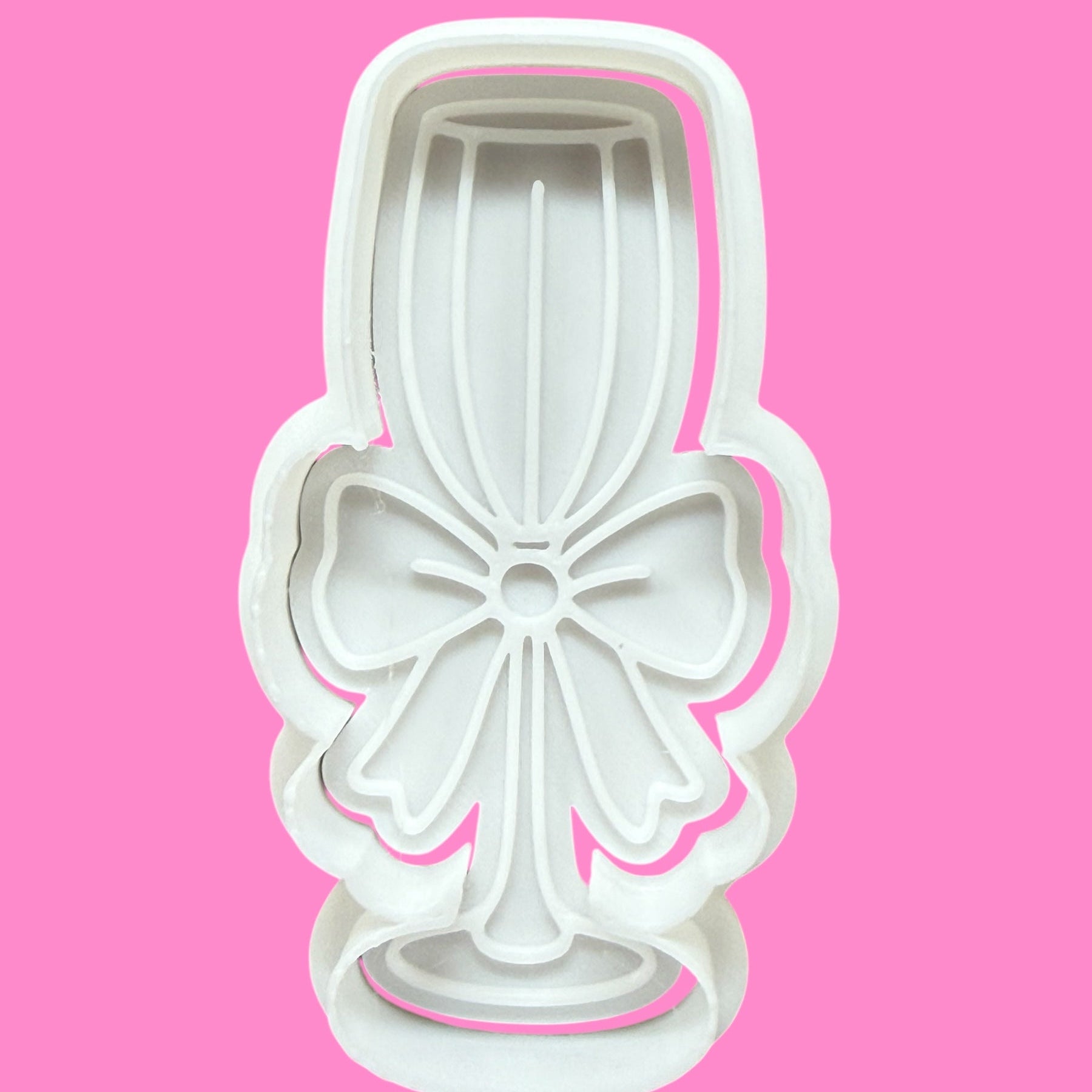 A white cookie cutter in the shape of a champagne glass with a bow detail on a pink background.