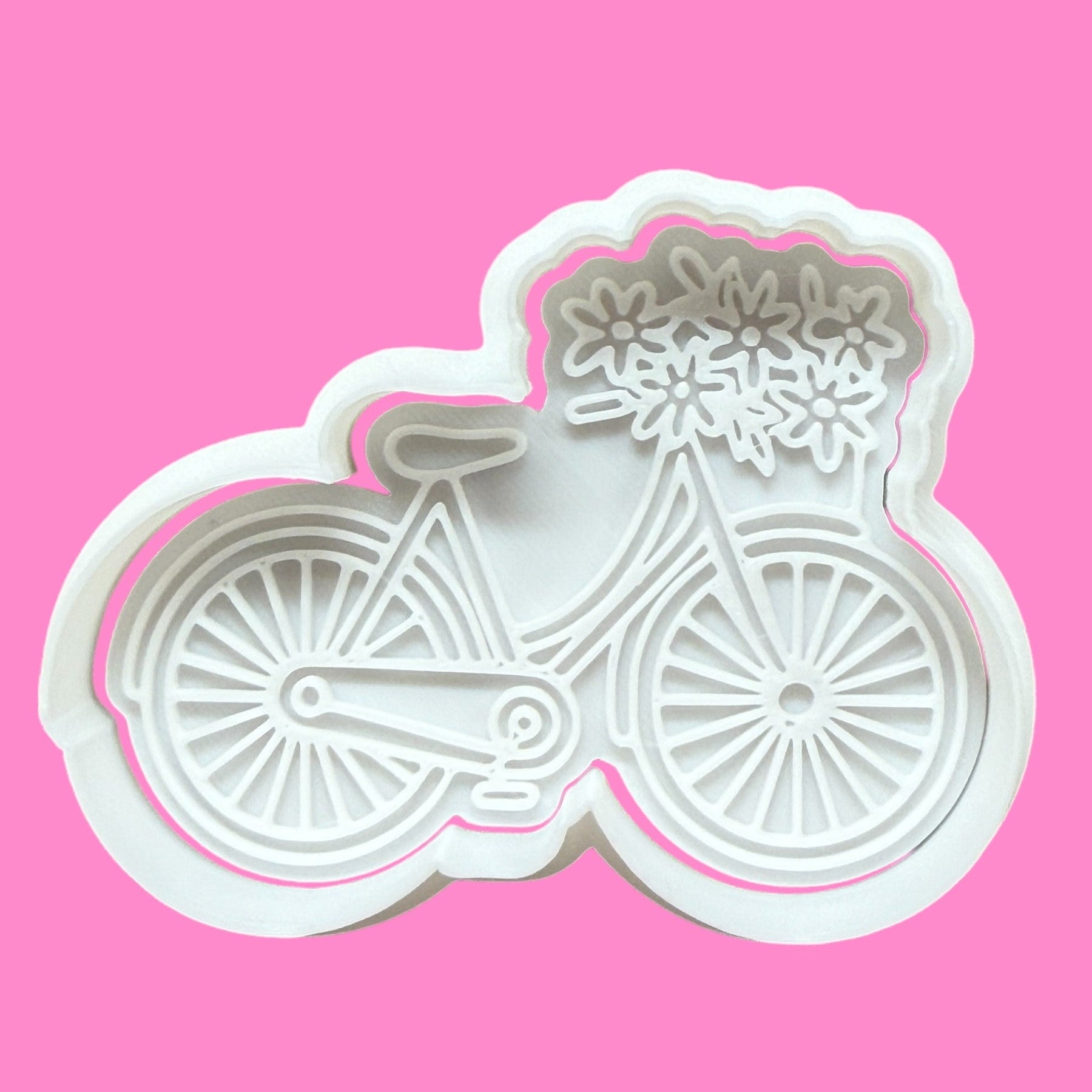 A white plastic cookie cutter and embosser in the shape of a vintage-style bicycle with a floral basket on top, displayed against a pink background.