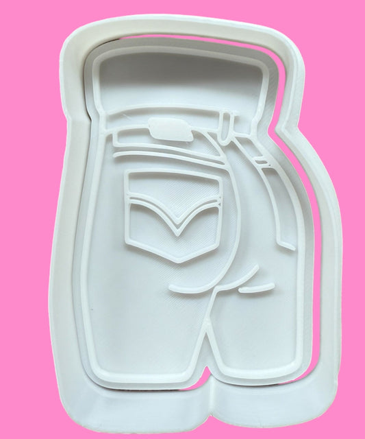 A white, denim-clad buttocks-shaped cookie cutter against a pink background.