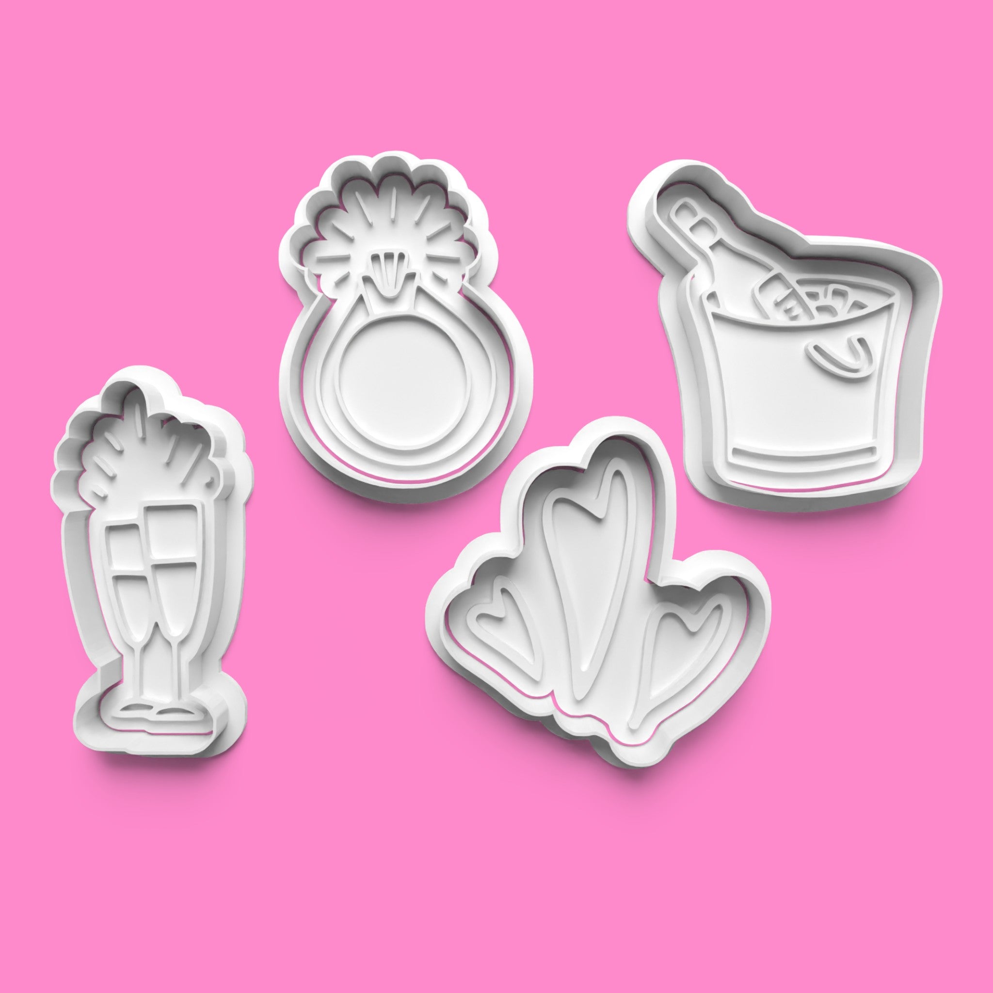 A set of white cookie cutters and stamps with engagement-themed designs, including rings, champagne glasses, and hearts, displayed on a pink background.