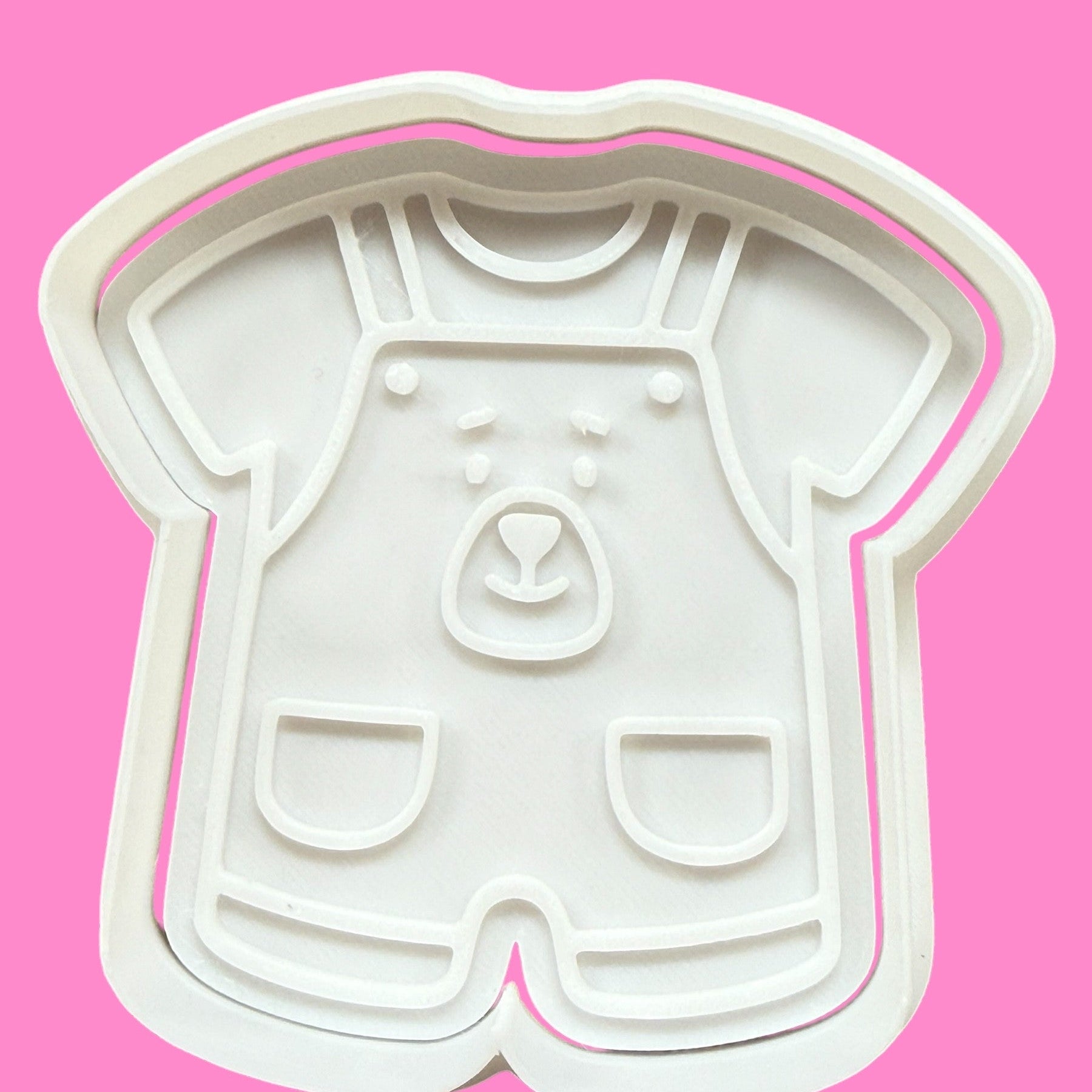 A white cookie cutter in the shape of a newborn romper with a bear face detail, displayed against a pink background.
