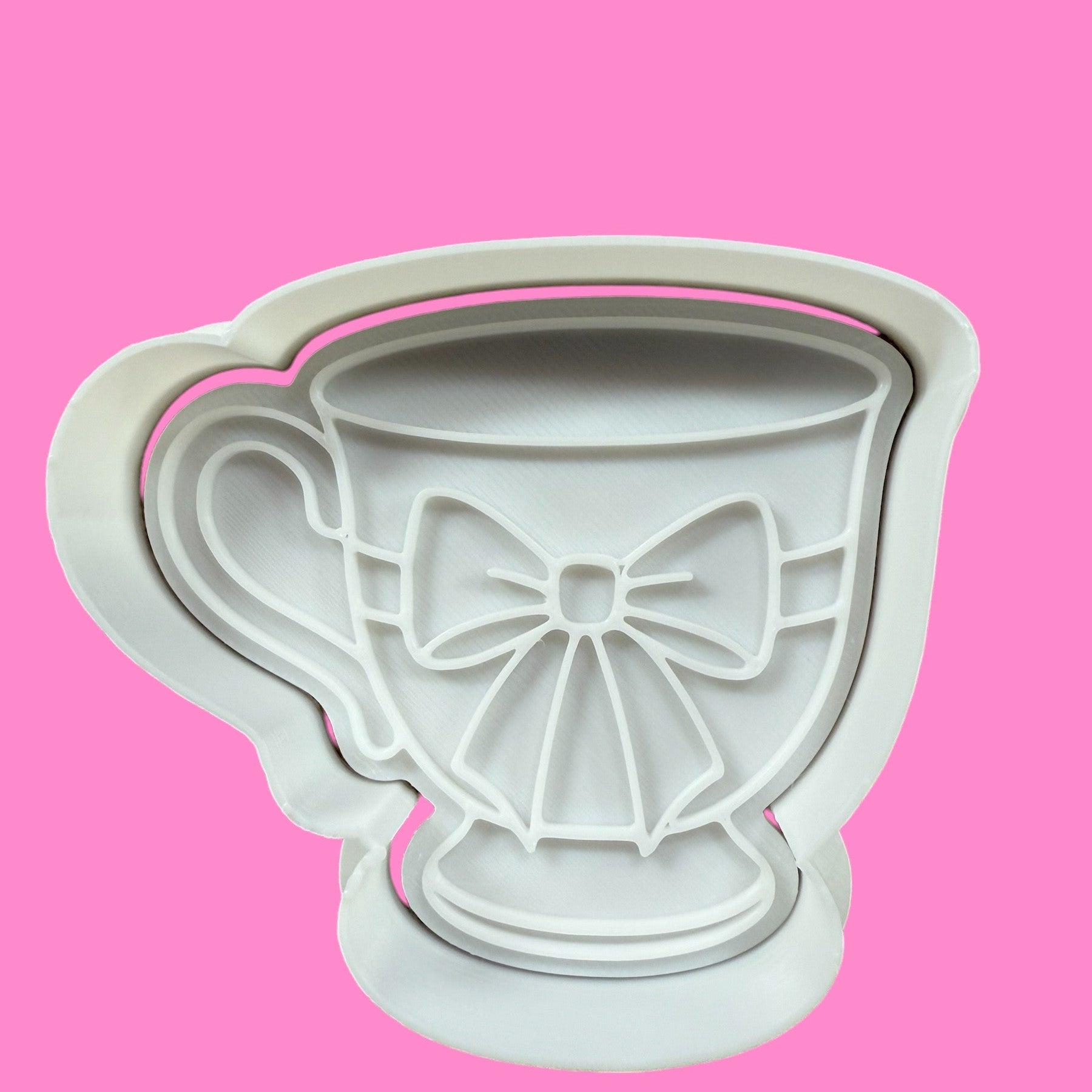 A white plastic cookie cutter and embosser in the shape of a teacup with a ribbon detail.