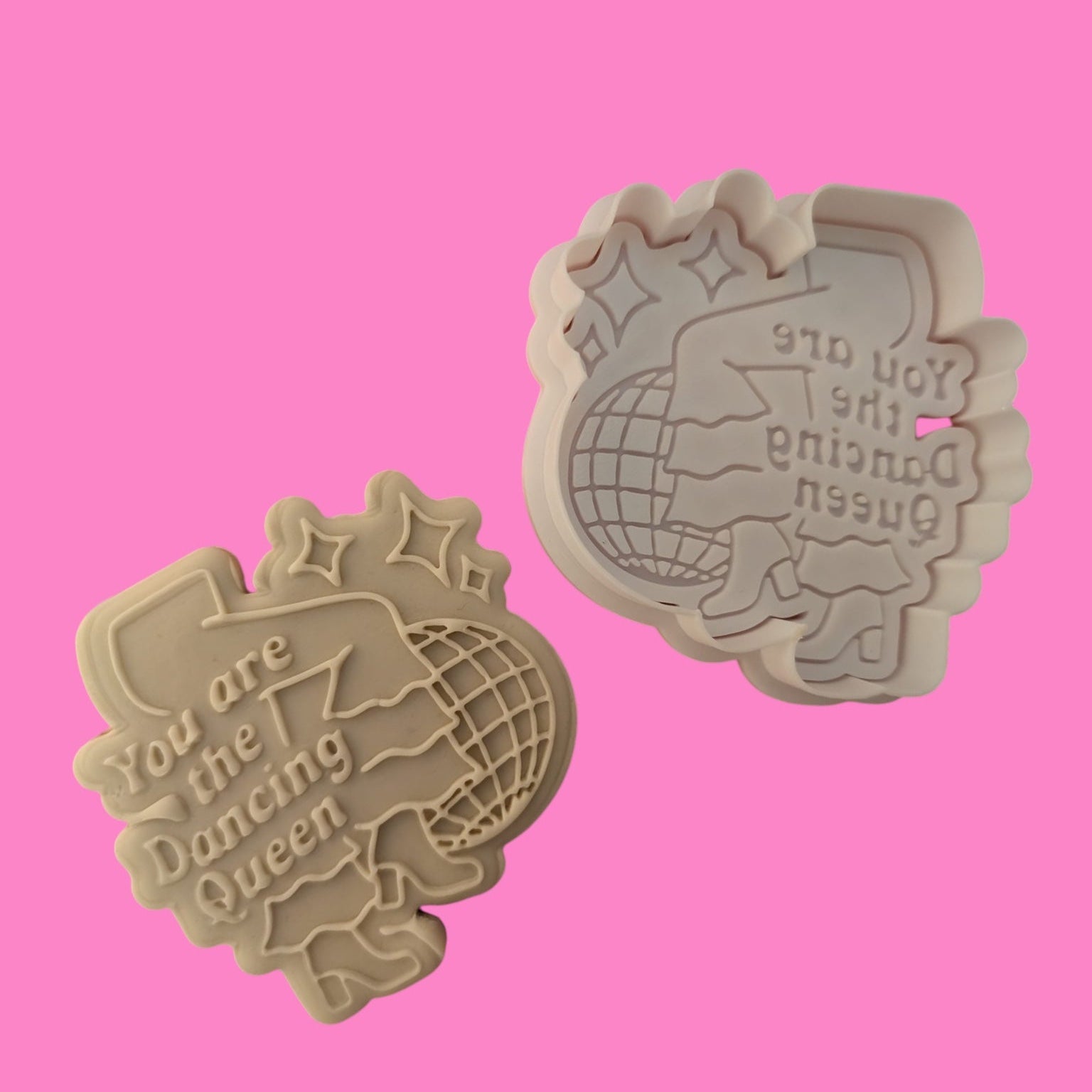 Two plastic cookie cutters with 'You Are the Dancing Queen' embossed design, one in a flat shape and the other with a raised pattern, displayed against a pink background.